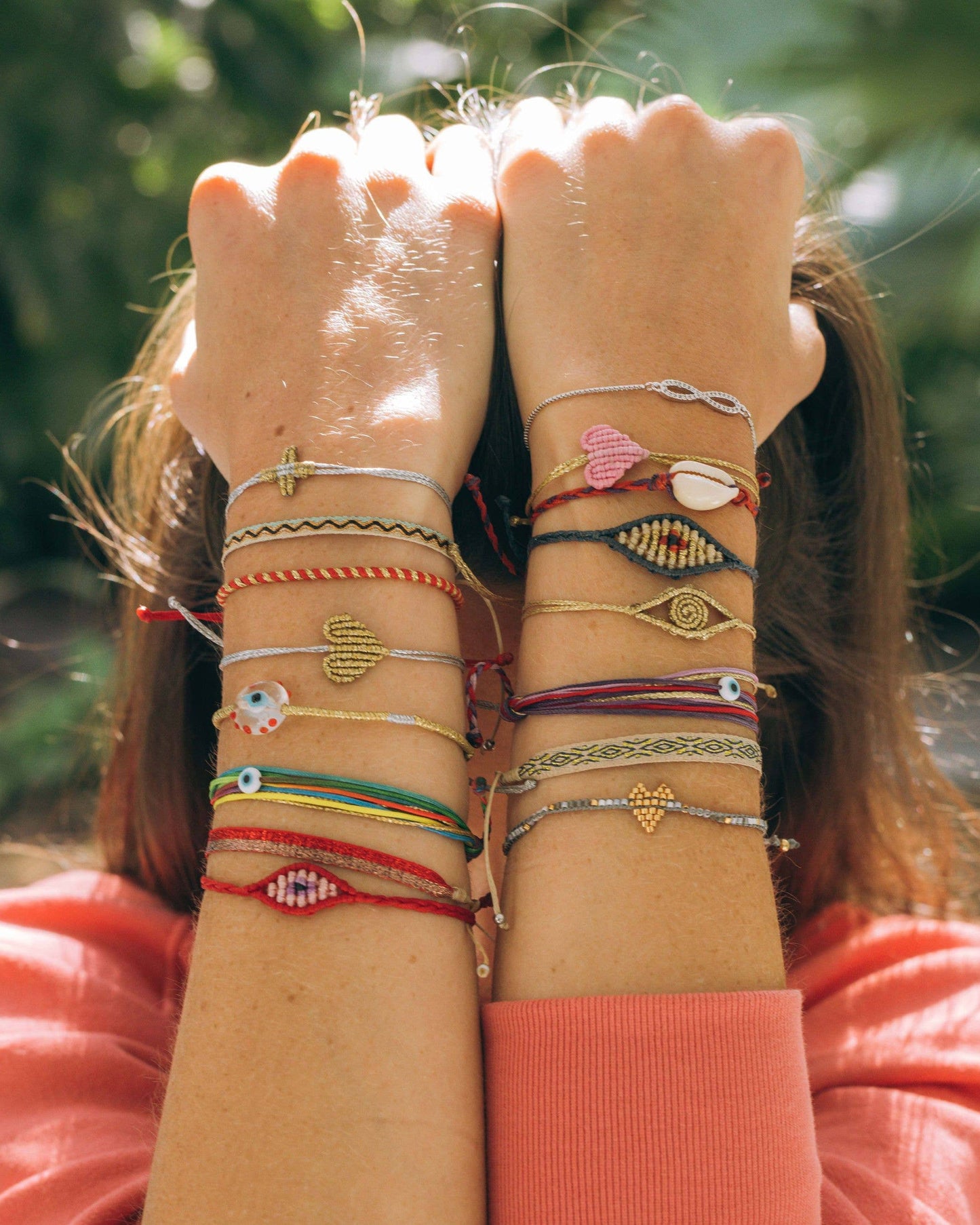 Person with both forearms raised, showing off multiple colorful handmade bracelets—including OIYA's beaded and charm designs, plus the OIYA TWIST Duo Color Evil Eye Bracelet—on each wrist; softly blurred greenery in the background.