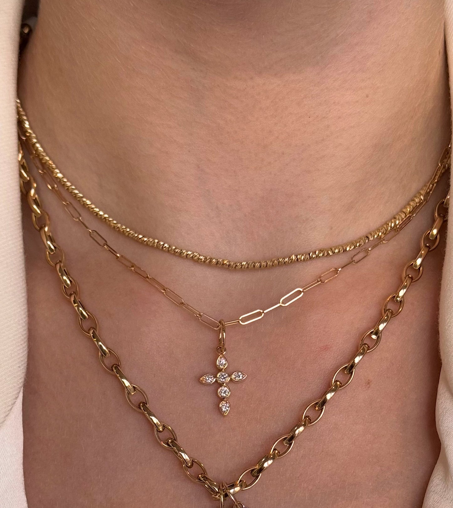 A close-up showcases a person in a cream top wearing three layered necklaces: the aira CISSY Gold Diamond Cross pendant, a yellow gold chain, and another with diamond-studded letters spelling "LOVED" vertically.