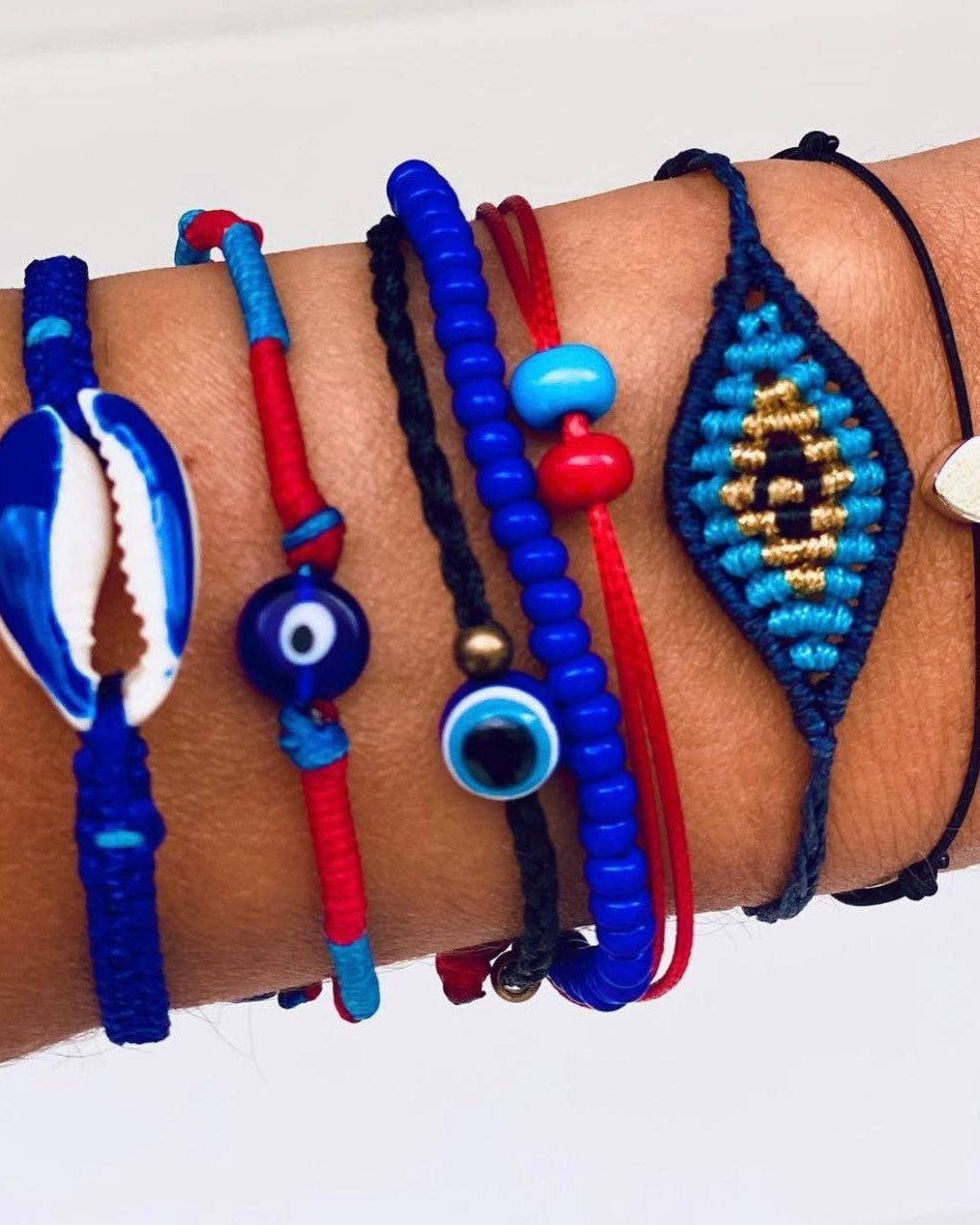 A wrist stacked with OIYA Alegria Bead Bracelets in blue and red, plus an evil eye charm, cowrie shell, and embroidered evil eye design by OIYA.