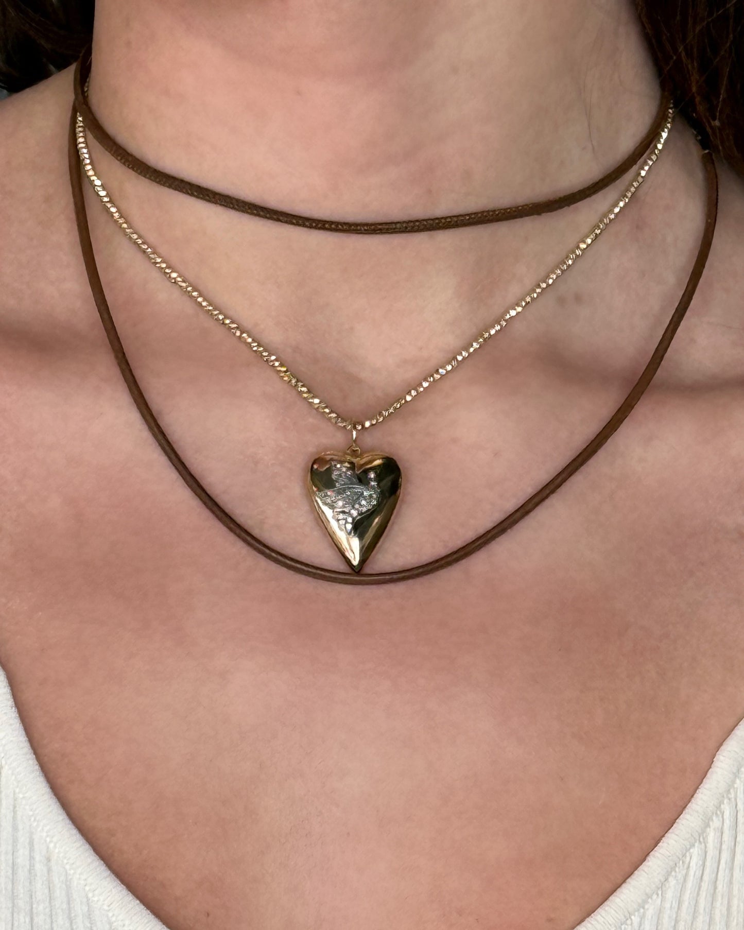 A person wears three brown and gold minimalist pieces, including the HANNAH Gold Leather Cord Necklace by Dylan James Jewelry/flower, featuring an adjustable metallic heart pendant over a white ribbed top.