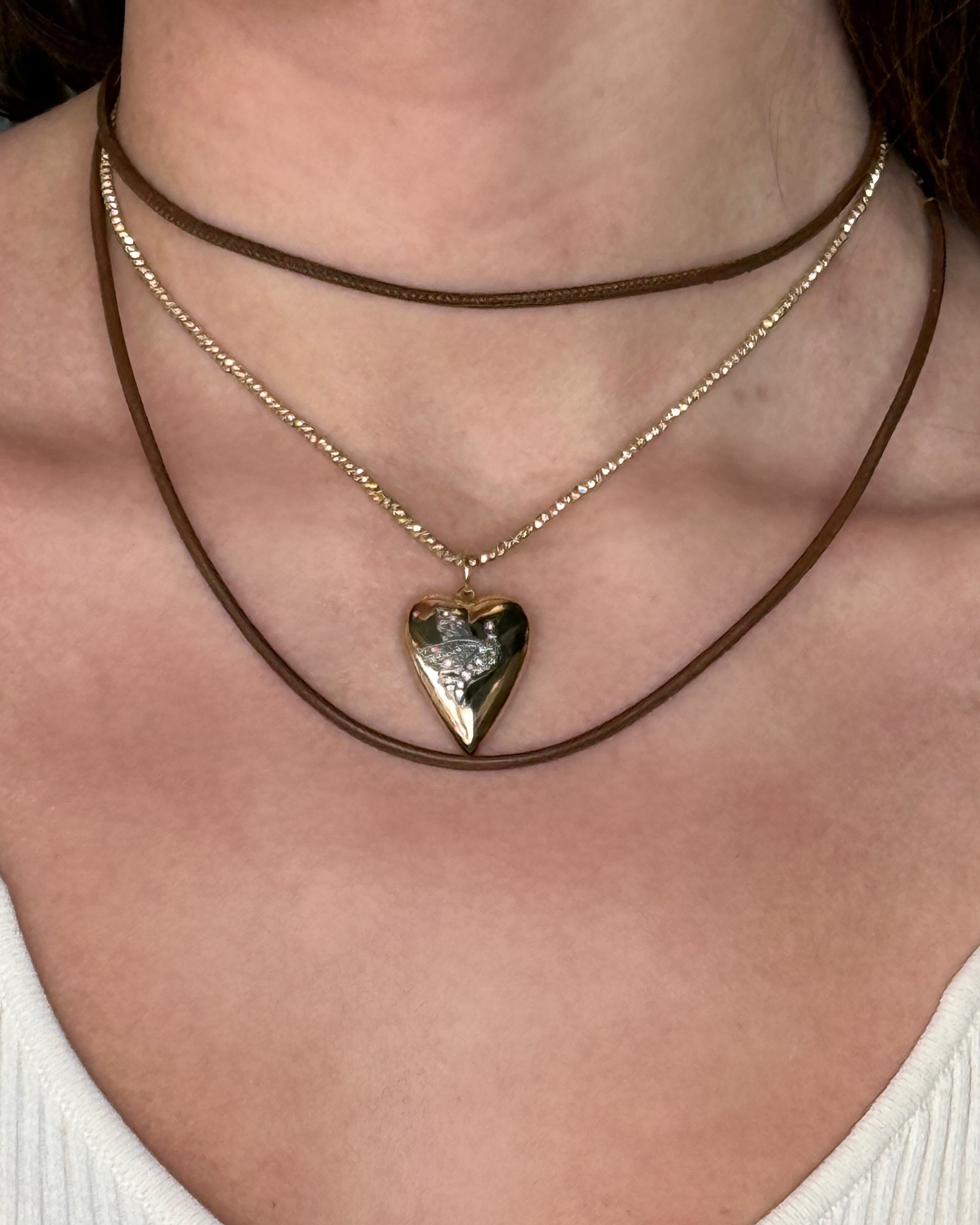 A person wears three brown and gold minimalist pieces, including the HANNAH Gold Leather Cord Necklace by Dylan James Jewelry/flower, featuring an adjustable metallic heart pendant over a white ribbed top.