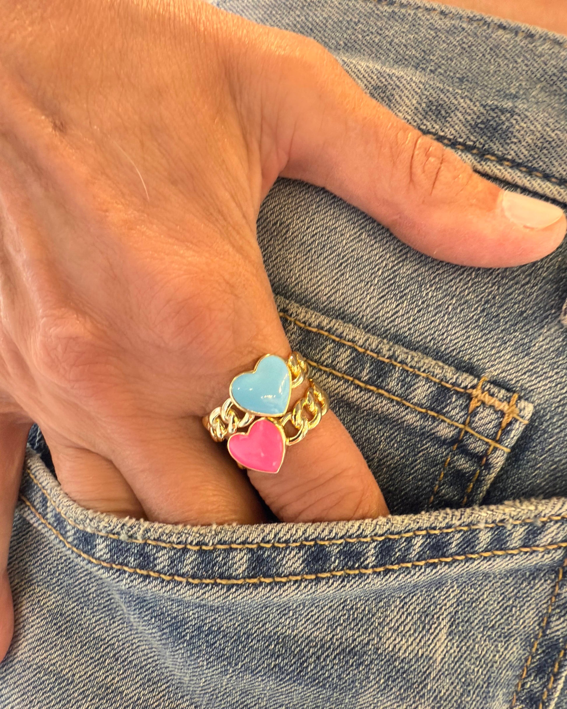 A hand with two Dylan James HAPPY Blue Heart Enamel Rings—gold plated silver bands with blue and pink hearts—rests on the edge of a denim jeans pocket.