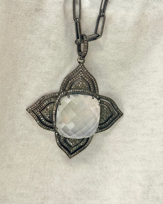The MELISSA Silver Pendant Necklace by Dylan James showcases a large, faceted diamond crystal in a quatrefoil shape, elegantly displayed on a light fabric background.