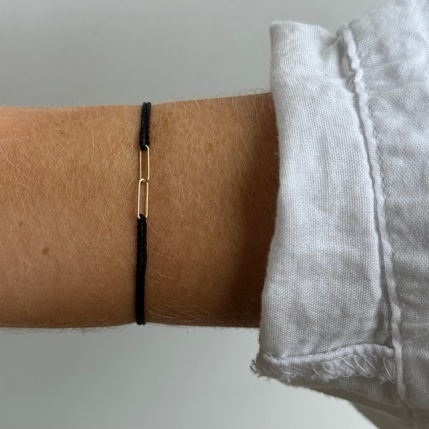 A person wearing the HORTENSE YOUNME Bracelet by hortense—a simple black adjustable cord with a small 14kt yellow gold rectangular link—styled on their wrist beside a white long-sleeve shirt for a modern jewelry touch.