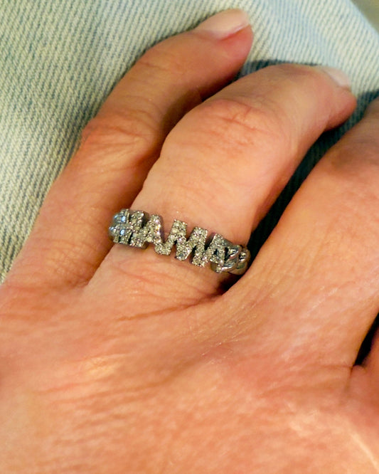A hand wearing the aira/djj MAMA Diamond Ring in oxidized sterling silver with "MAMA" lettering, set against a light blue fabric background.