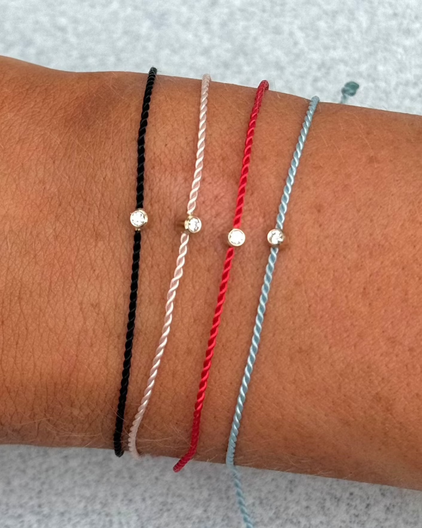 A wrist adorned with four thin braided bracelets in black, white, red, and blue, each featuring a small bead at the center. Among them, the SOL Gold Cord Bracelet by hortense stands out with its adjustable design and gleaming 14kt yellow gold charm.