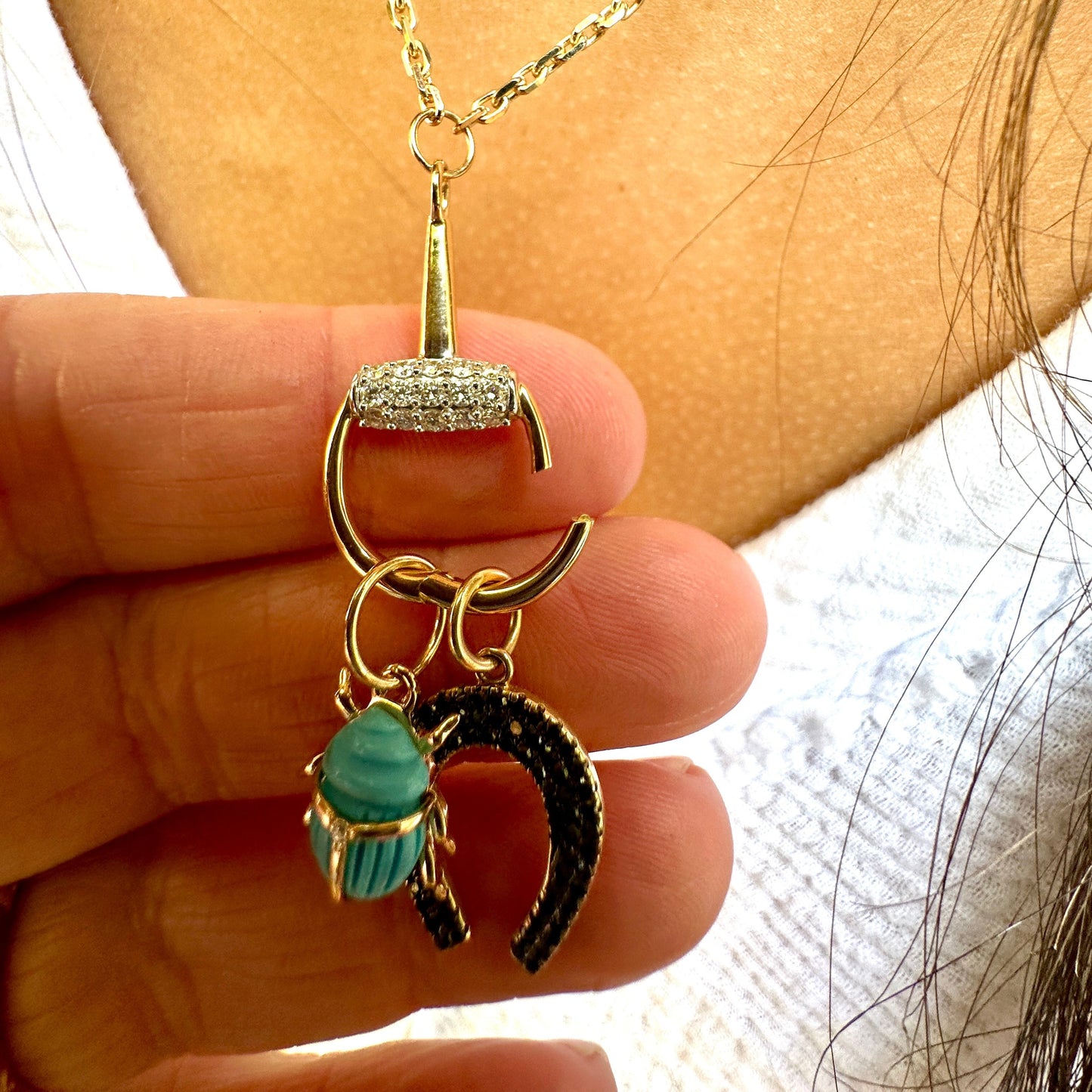 A person holds the BUD Gold Diamond Necklace by AIRA featuring horse bit charms with a turquoise gem, pavé diamond bar, and black rhinestone crescent, all set in 14kt yellow gold.