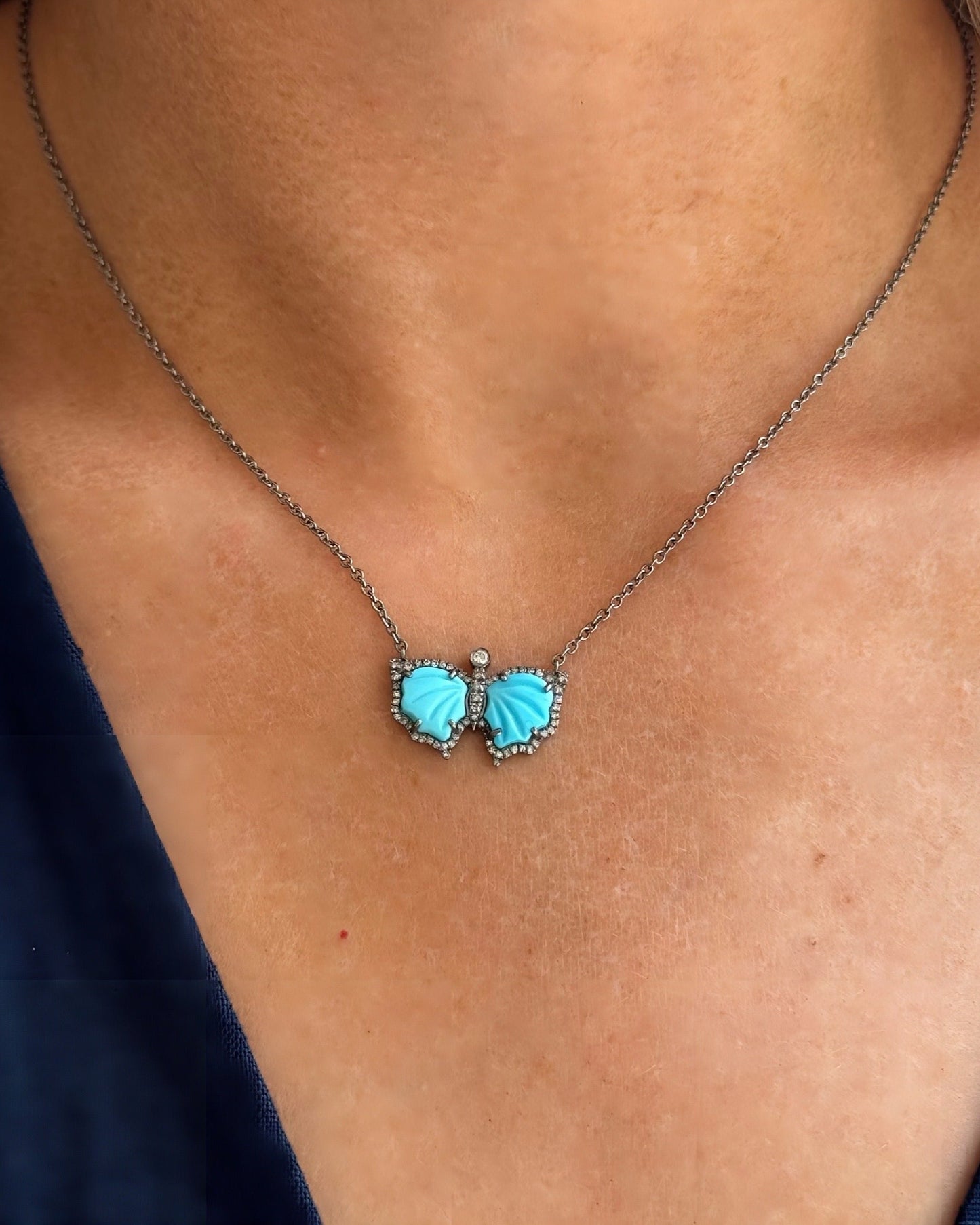 The BLAIR Butterfly Necklace by DYLANJAMES features a silver chain and turquoise accents, its vibrant pendant resting elegantly against light skin.