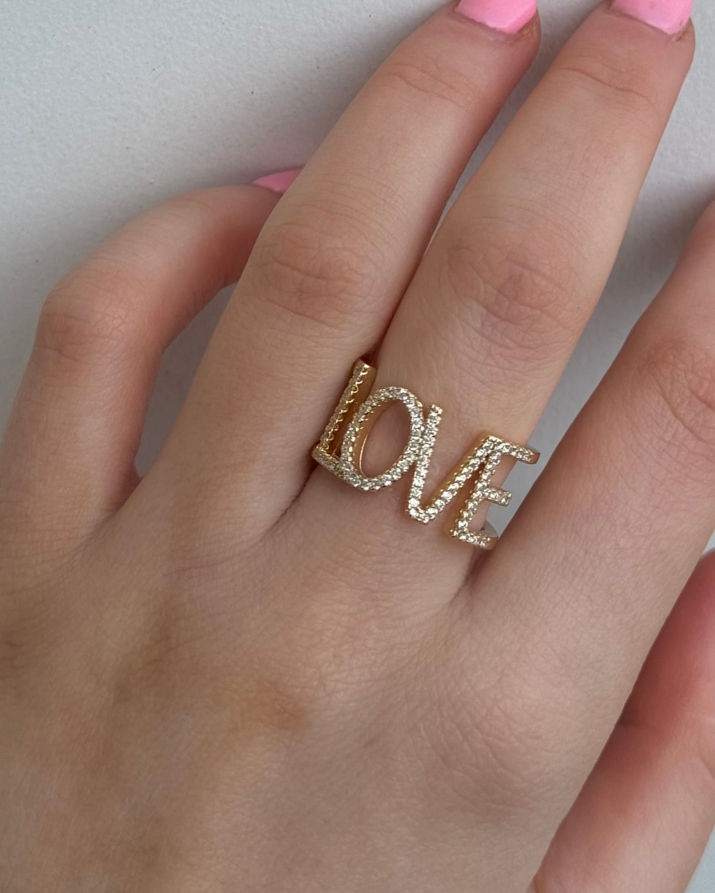 A hand with pink nails displays the Dylan James Jewelry LOVE Gold Diamond Ring, elegantly shimmering with a touch of cubic zirconia.