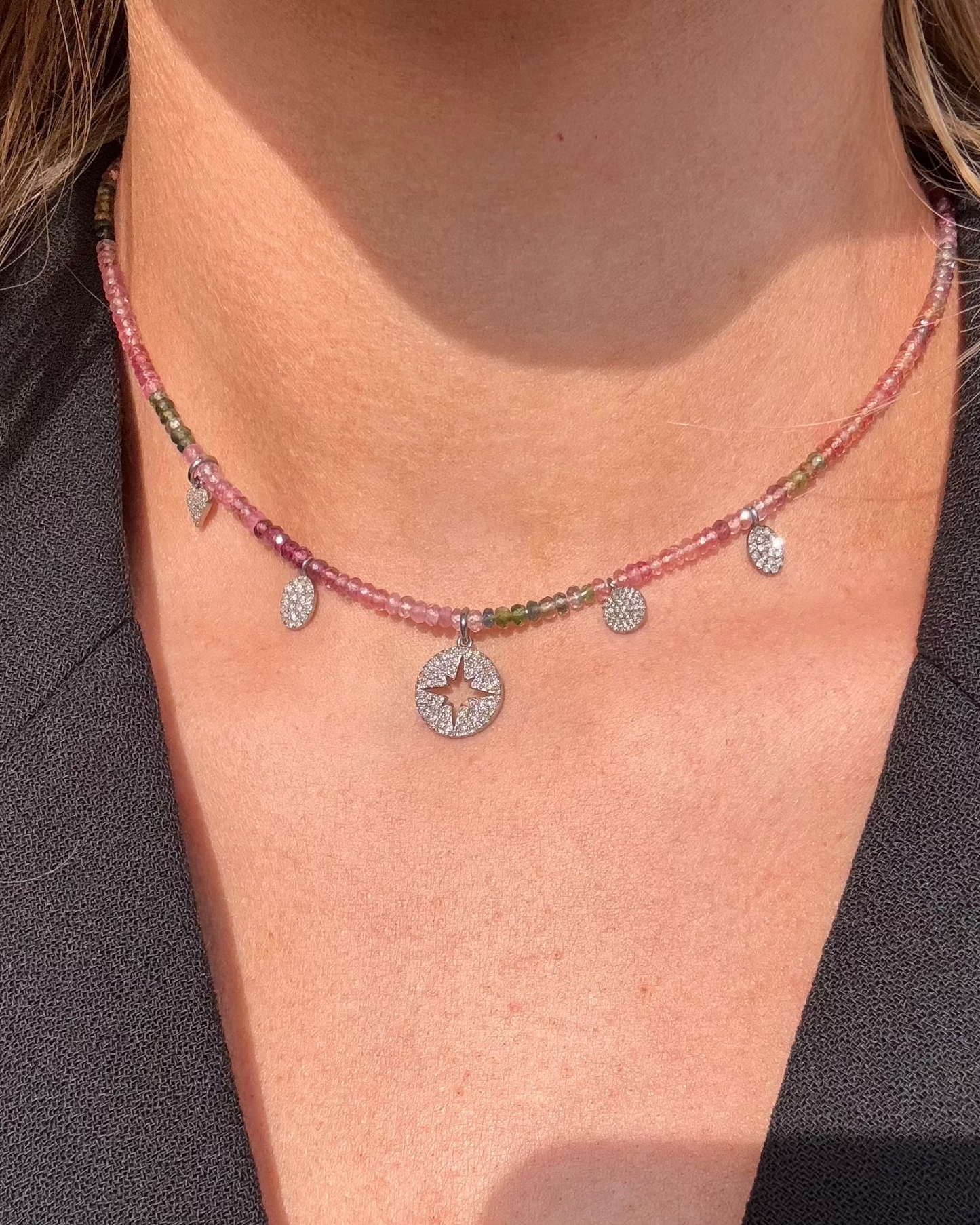 A woman showcases the MATHIAS Tourmaline Diamond Charm Necklace by Dylan James Jewelry, a multi-colored creation with tourmaline beads and circular and teardrop pendants, elegantly set against a black top.