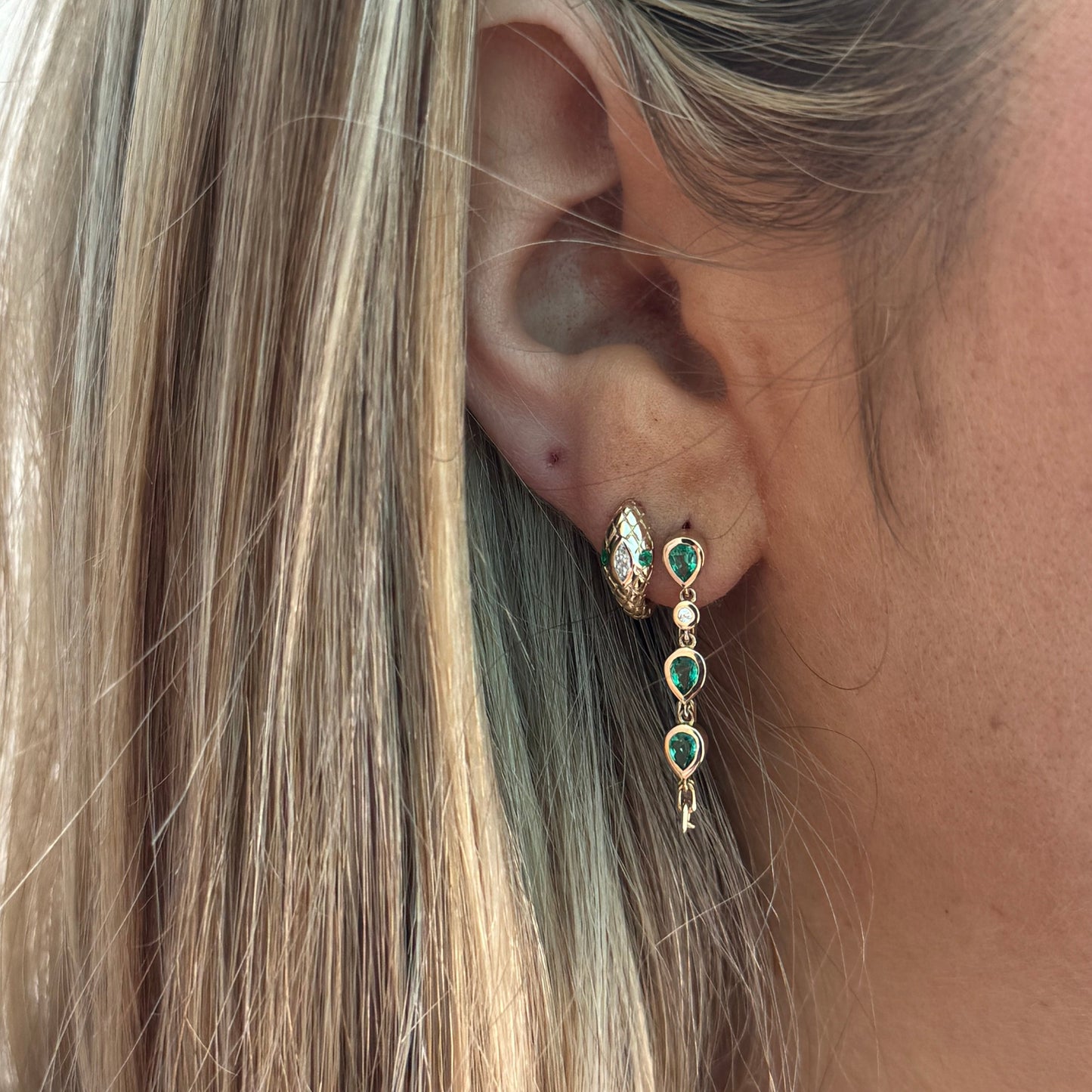 Close-up of a woman's ear wearing two gold earrings: a small 14kt yellow gold hoop and the BELLA Emerald Earring by Dylan James Jewelry—featuring green stones with delicate gold accents.