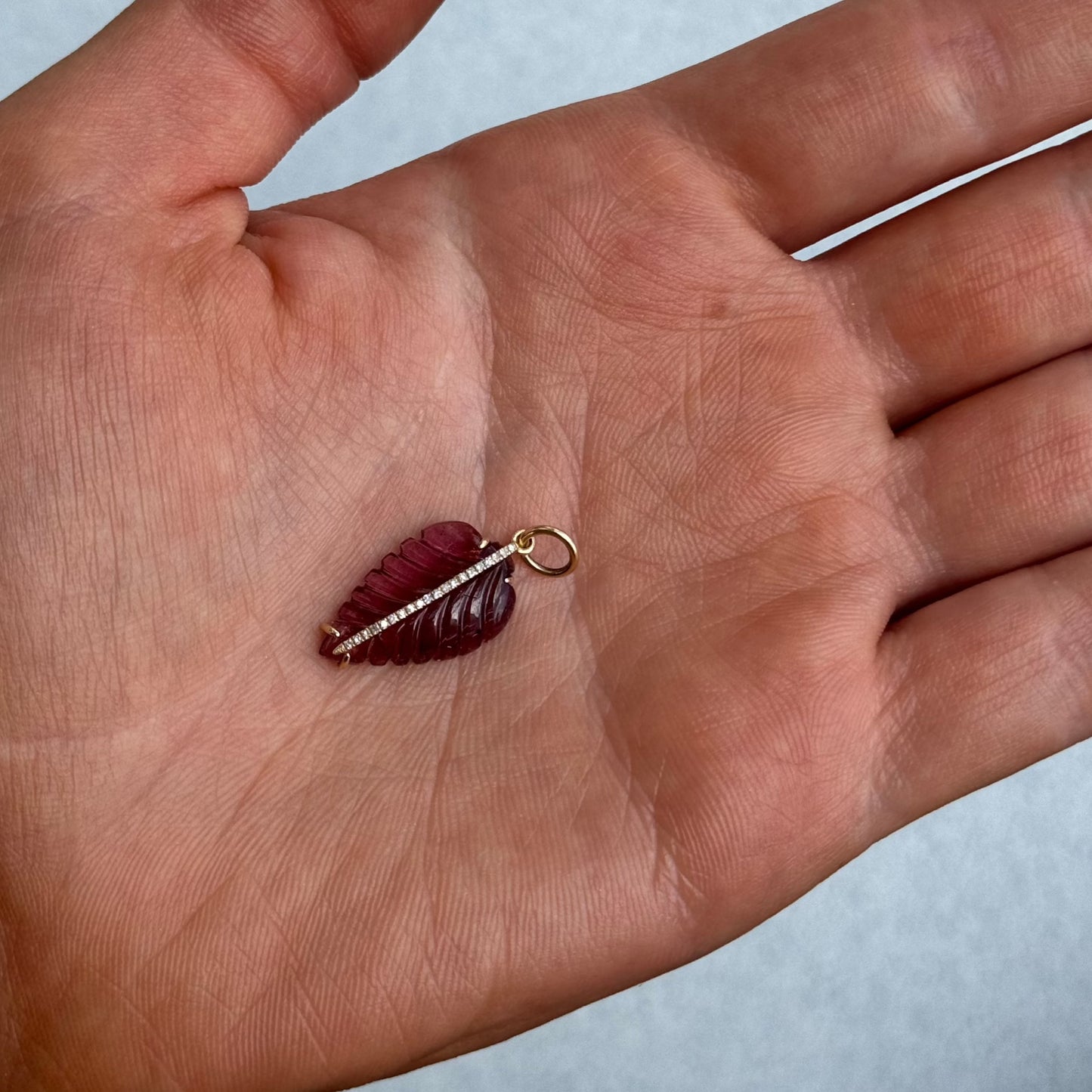A hand holds the LEAF Charm by Dylan James Jewelry—a small, red, leaf-shaped pendant in 14kt yellow gold, featuring a gold loop and a row of tiny white stones along its center.