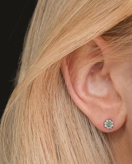 Close-up of an ear with straight blond hair, wearing a small flower-shaped earring crafted from silver, adorned with emerald-green and clear stones: the MARGEAUX Emerald Studs by Dylan James Jewelry.
