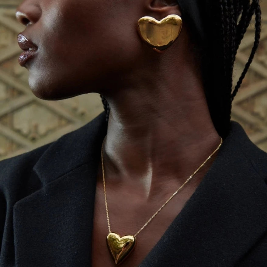 A person wearing PUFFY Heart Earrings by Dylan James Jewelry and a matching gold necklace with a dark jacket. The statement earrings catch the light beautifully against the geometric patterned background.