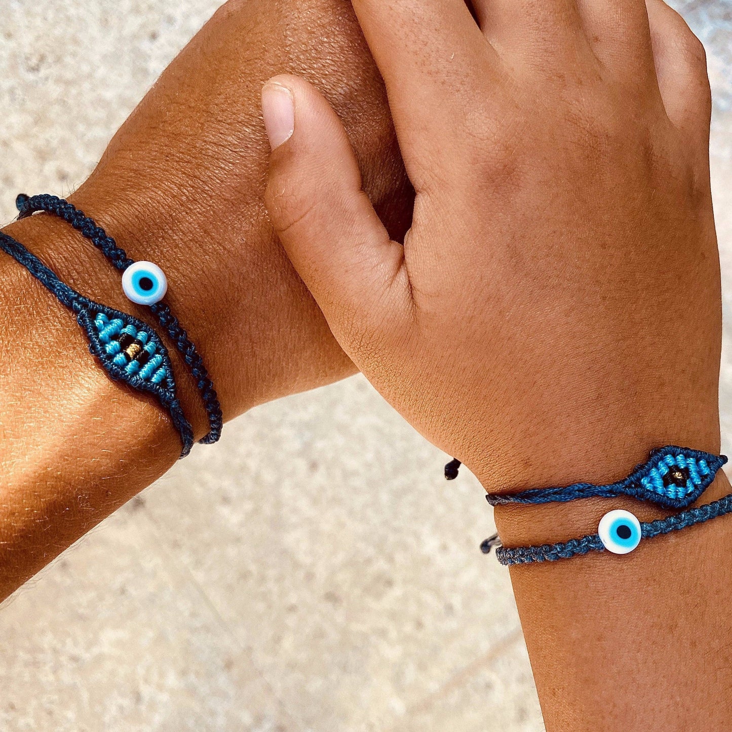 Two hands, against a neutral background, wear matching OIYA Gold Evil Eye Braided Bead Bracelets. These adjustable handmade bracelets by OIYA feature intricate beadwork and meaningful evil eye charms for a stylish accessory.