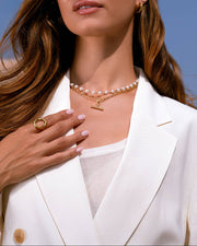 A woman with long brown hair wearing a white blazer, a pearl necklace with a gold T-shaped pendant, and gold hoop earrings, set against a clear blue sky.