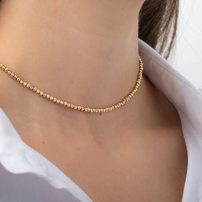 A close-up showcases the SPARK Gold Necklace by Dylan James Jewelry, a 14kt yellow gold creation featuring diamond cut beads, elegantly draped over a woman's neck and paired with a crisp white collared shirt.