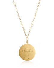 A gold paperclip chain necklace with a round brushed gold pendant engraved with the text 'BALANCED AF' and 'JLA', shown on a clean white background.