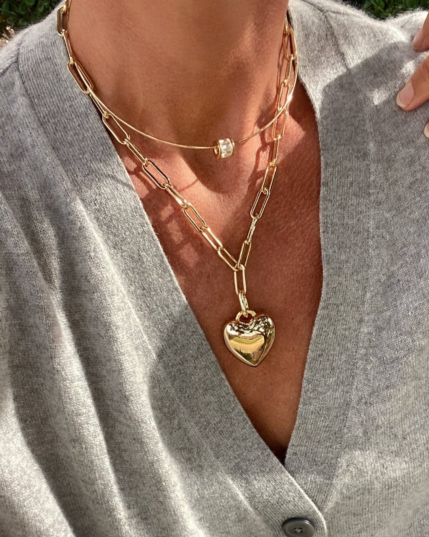 Close-up of a person wearing a gold chain necklace with a heart pendant on a gray shirt.