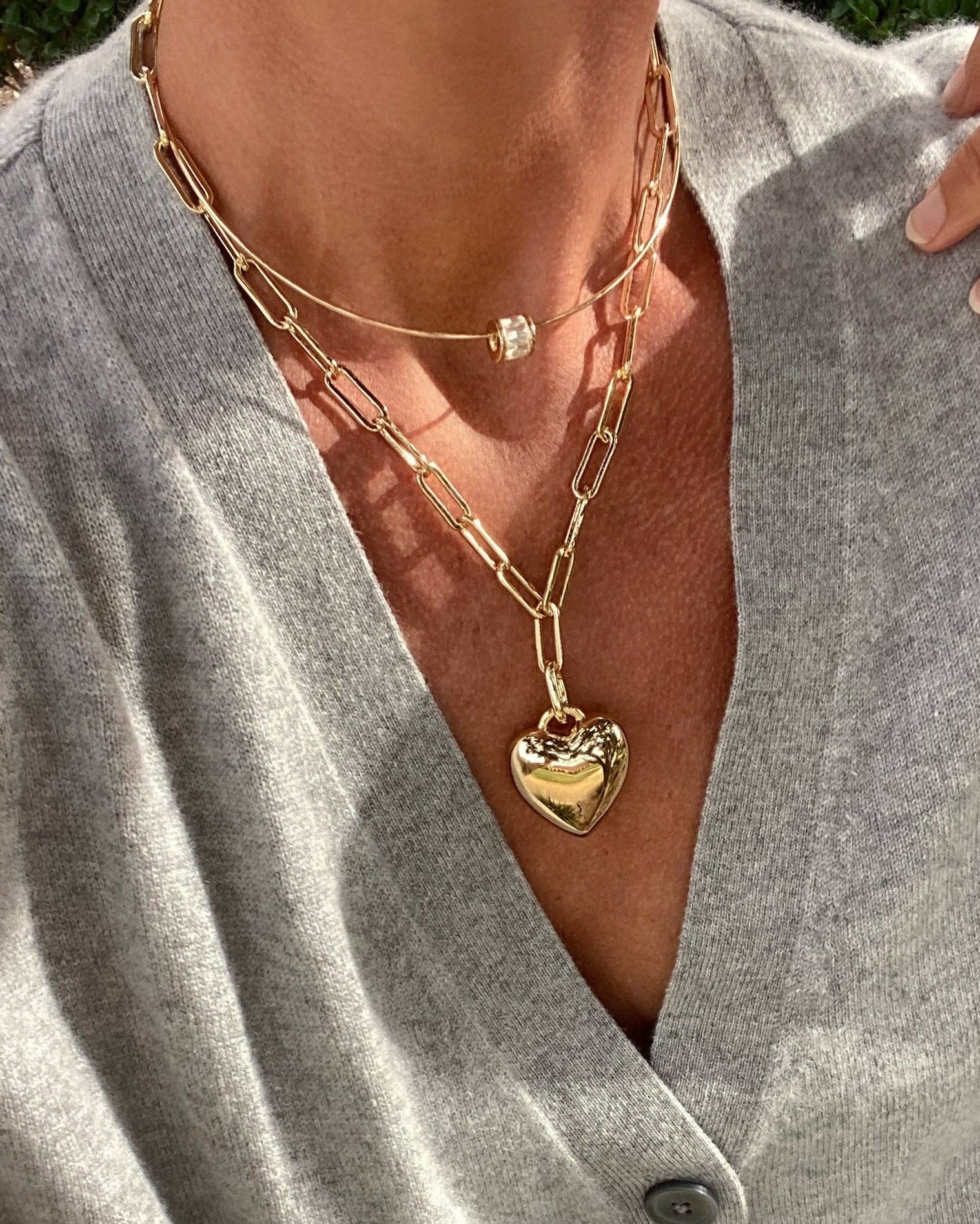 Close-up of a person wearing a gold chain necklace with a heart pendant on a gray shirt.