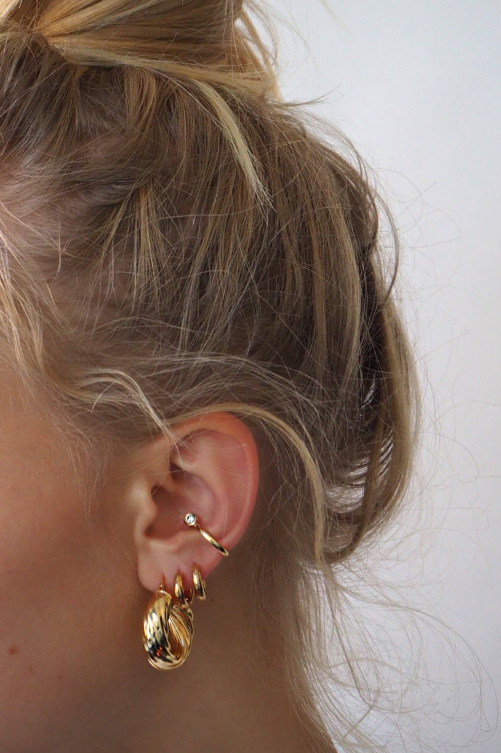 Close-up of a person's ear showcasing a chic silver stud, the elegant Lili Claspe REESE Conch Ear Cuff in gold-plated brass, and three stylish gold hoop earrings; hair is blonde and styled in a loose bun.