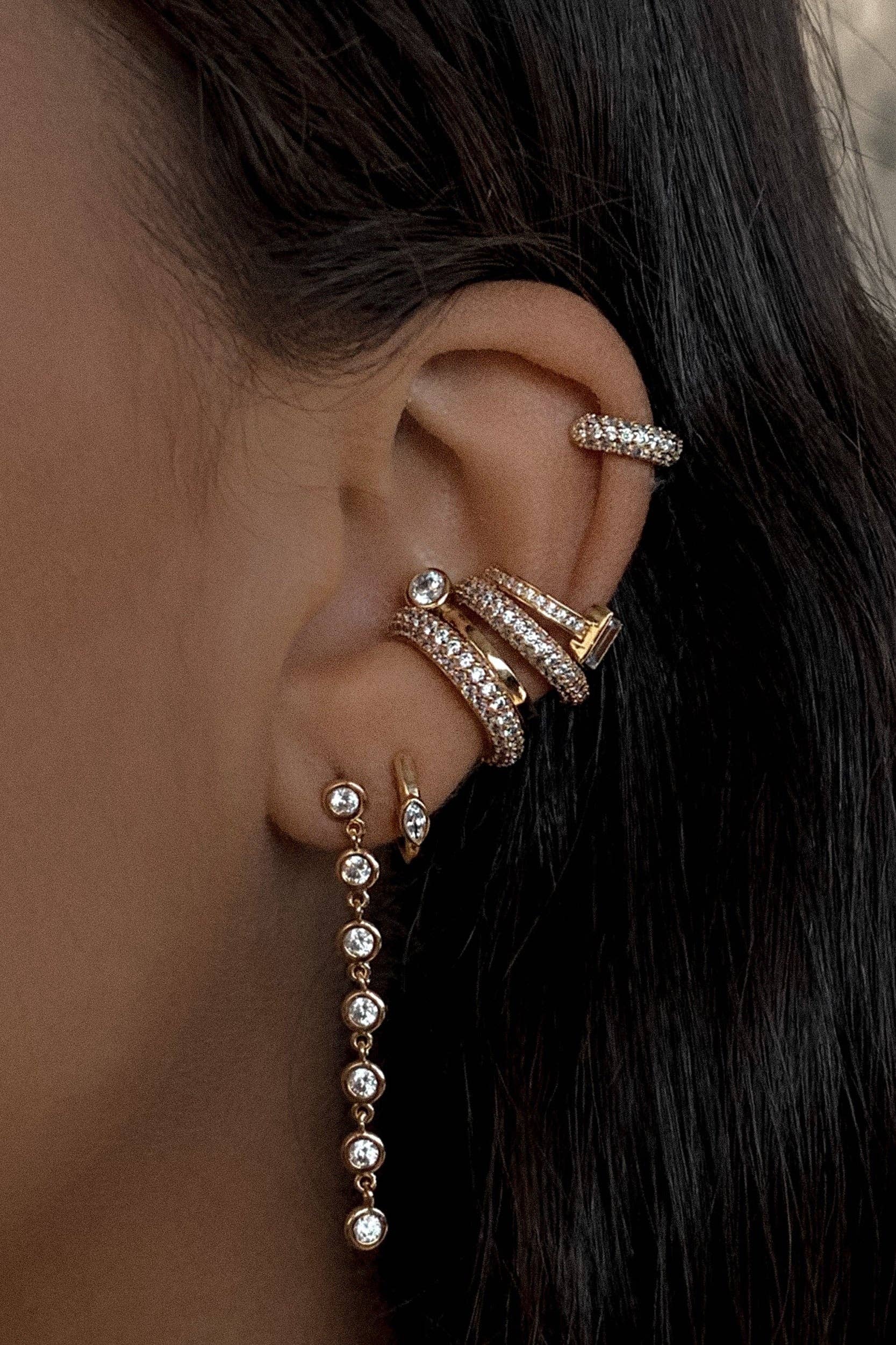 A close-up of an ear adorned with multiple gold hoop earrings and a dangling earring with small gemstones, complemented by the chic REESE Conch Ear Cuff by Lili Claspe.