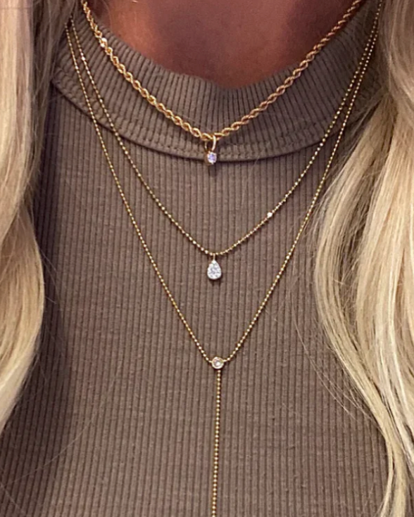 Layered gold necklaces on a person wearing a brown ribbed sweater with blonde hair.