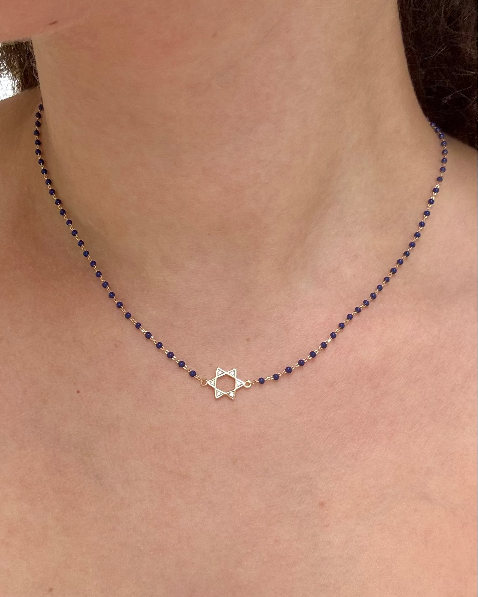 A delicate necklace worn on a person's neck, featuring a small silver Star of David pendant at the center, set against a dark beaded chain.