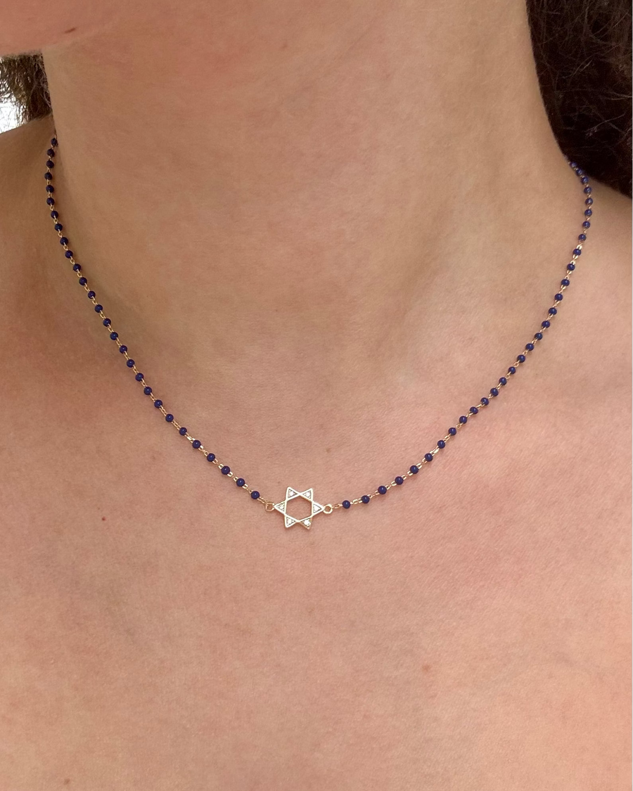 A delicate necklace worn on a person's neck, featuring a small silver Star of David pendant at the center, set against a dark beaded chain.