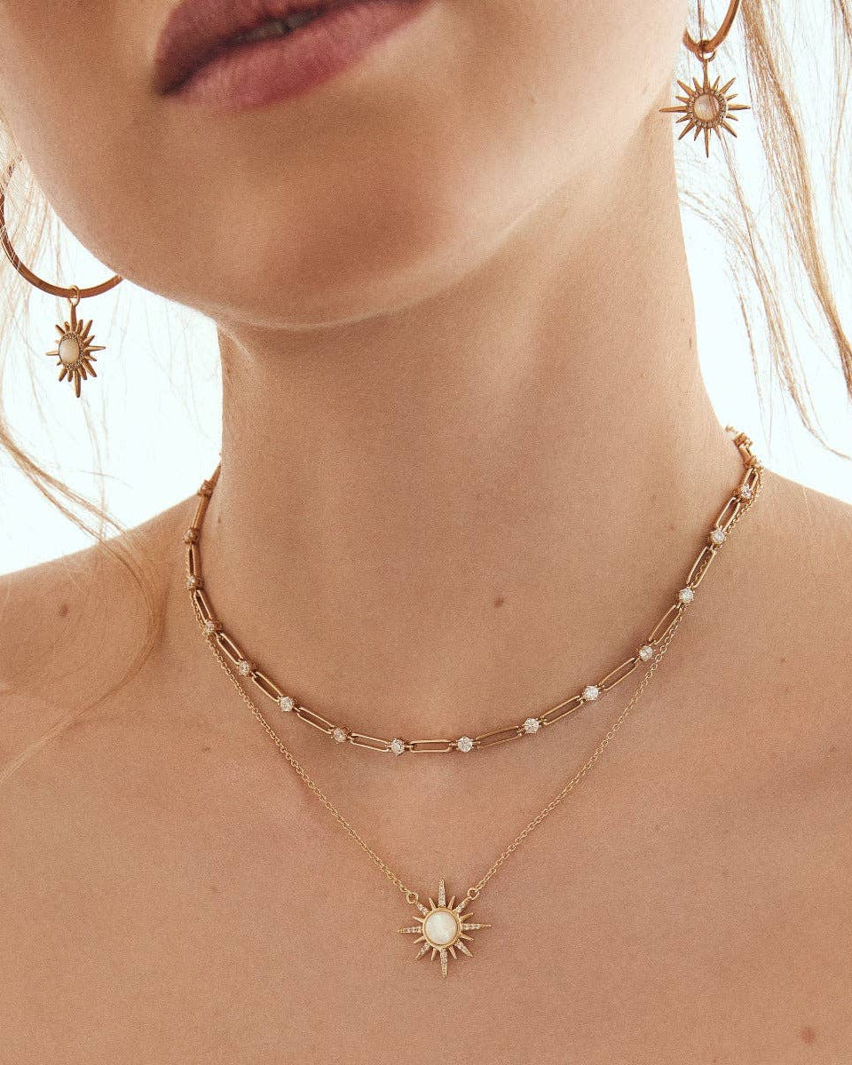 A woman wears Elizabeth Stone Jewelry’s ELIZABETH STONE Gem Starburst Necklace layered with a 14k gold-plated necklace, gold sun-shaped earrings, and a matching sun pendant.