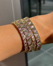 A close-up of a gold-toned multi-band cuff bracelet with pyramid-shaped crystal-encrusted panels, worn on a person's wrist.