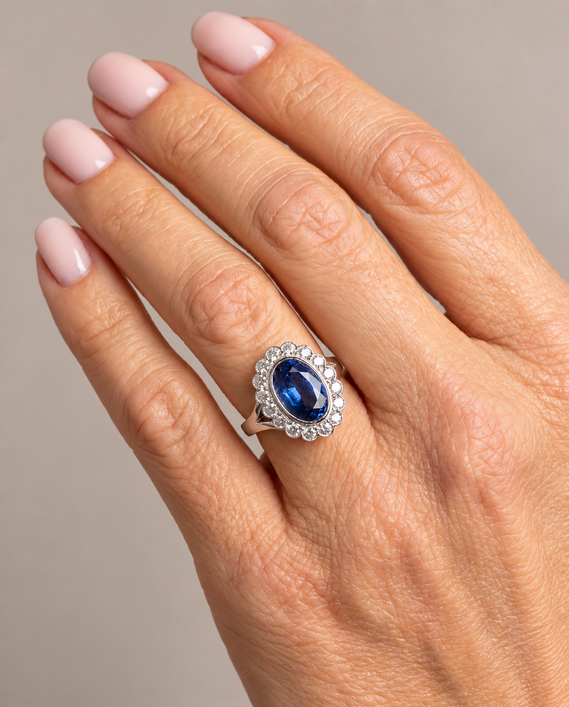 A close-up of a hand wearing a silver ring with an oval blue sapphire center stone and a halo of small clear stones, set against a neutral gray background.
