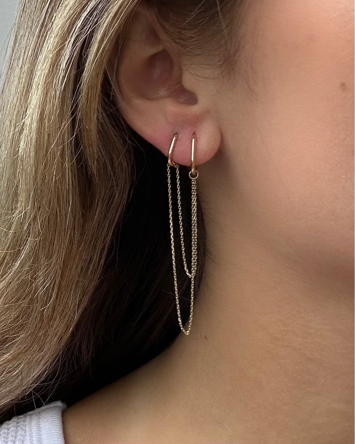 Close-up of a person wearing the Métier By Tomfoolery Double Add-on Chain by Métier by Tomfoolery, featuring double hoop earrings joined by two gold chains—the ideal Hoop Earring Add-On for a stylish layered look.