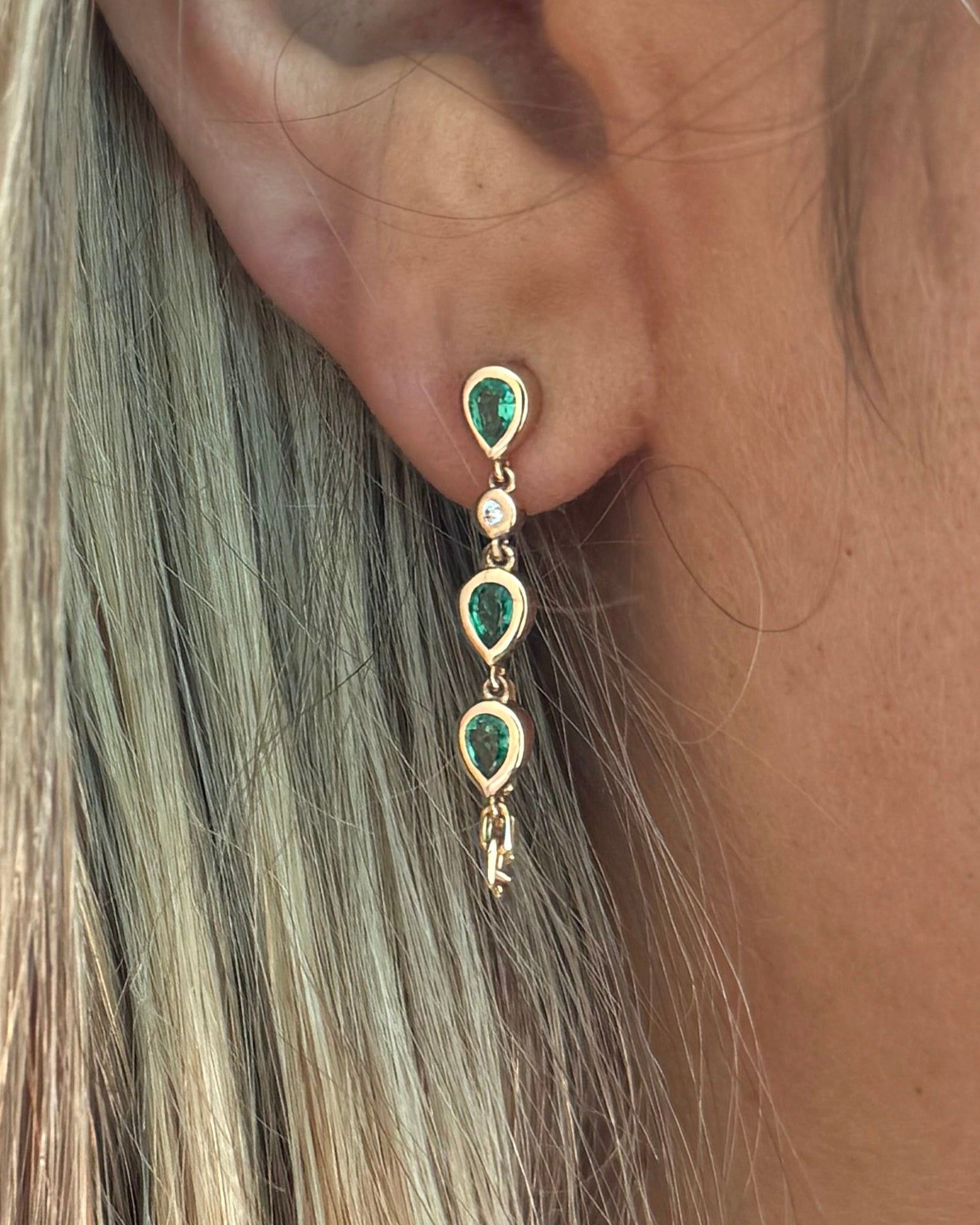 Close-up of a person's ear wearing the BELLA Emerald Earring by Dylan James Jewelry, crafted in 14kt yellow gold with three green teardrop stones and a small round white stone in a vertical design.