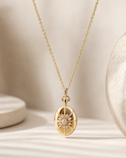 A gold pendant necklace featuring a compass rose design with a central cluster of small clear stones, displayed on a soft beige surface with a textured ceramic object in the background.