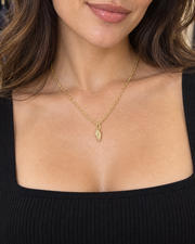 A woman with wavy brown hair wearing a delicate gold chain necklace with a small leaf-shaped pendant, paired with a black ribbed tank top in a retail store setting.