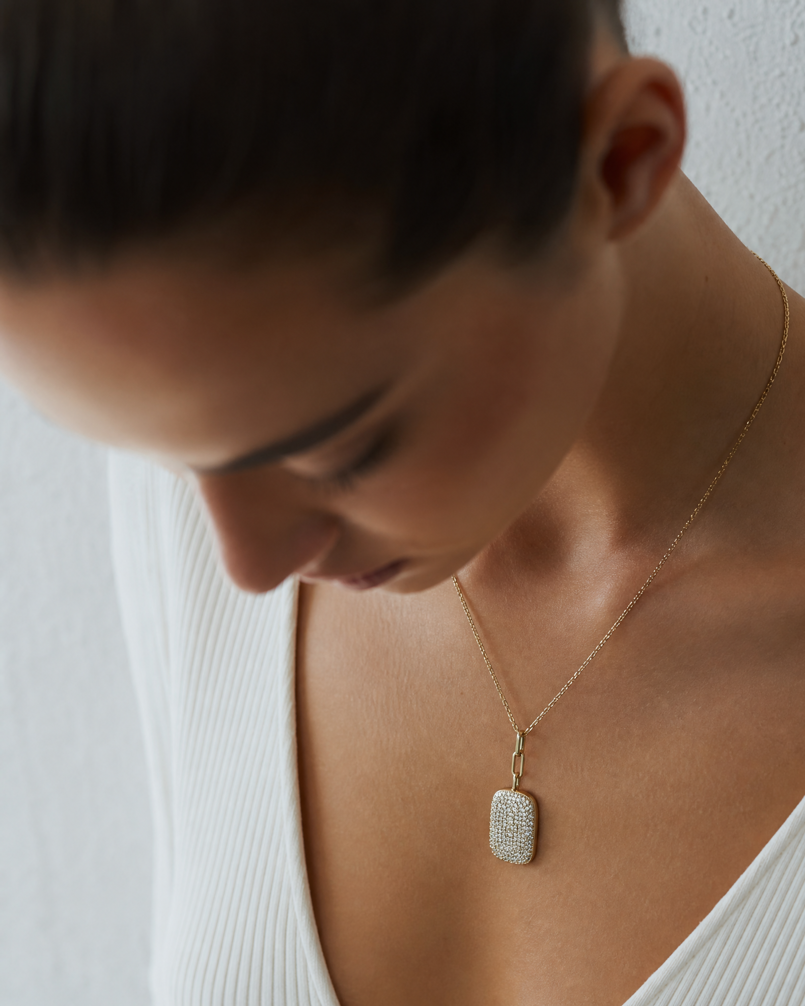 A close-up lifestyle shot of a woman wearing a delicate gold chain necklace with a textured, rectangular pavé pendant. She is wearing a white ribbed top against a neutral, light-colored textured wall.
