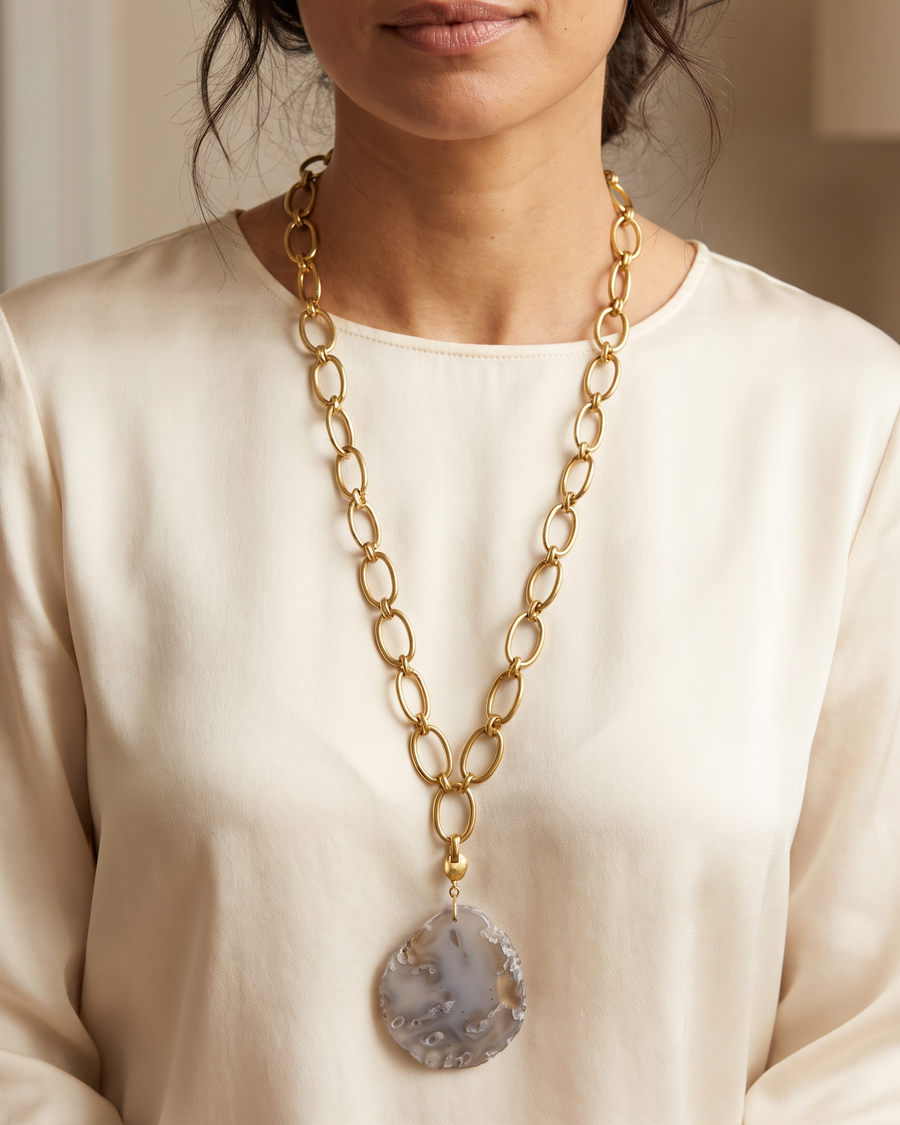 A woman wearing a long gold chain necklace with a large teardrop-shaped agate pendant, paired with a cream-colored long-sleeve top.
