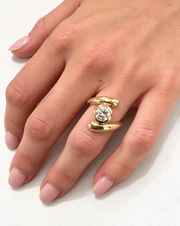 A close-up of a hand wearing a gold ring with a round clear gemstone. The ring features a textured gold band with a central bezel-set round diamond.