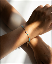 A delicate gold chain bracelet with small turquoise blue beads and a tiny central charm, shown worn on a person's wrist against a bright white background with strong natural shadows.