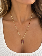 A close-up of a model wearing a delicate gold chain necklace with a small, textured reddish-brown feather pendant.