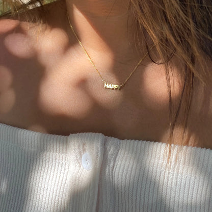 A person wearing an off-shoulder white top and a yellow gold plated "HAPPY" pendant from the HAPPY Gold Necklace by Thatch rests elegantly on their 14" chain, while shadows of leaves gently cascade across the skin.