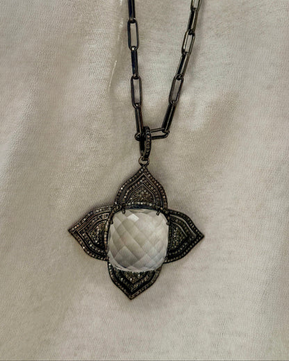 The MELISSA Silver Pendant Necklace by Dylan James showcases a large faceted Diamond Crystal in a decorative four-point frame on a textured 30" chain, elegantly set against a light fabric background.