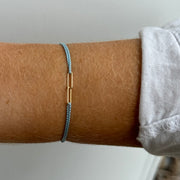 A close-up of a wrist wearing the hortense HORTENSE YOUNME Bracelet—a simple blue adjustable cord with a small 14kt yellow gold bar—paired with a white long-sleeve shirt for a sleek, modern look.
