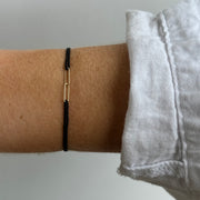 A person wearing the HORTENSE YOUNME Bracelet by hortense—a simple black adjustable cord with a small 14kt yellow gold rectangular link—styled on their wrist beside a white long-sleeve shirt for a modern jewelry touch.