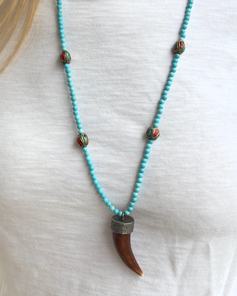 The JOE turquoise Beaded Necklace by Dylan James Jewelry features red and green inlay accent beads and a bold horn-shaped pendant, perfect for styling over a white shirt.