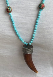 The JOE turquoise Beaded Necklace by Dylan James Jewelry features inlay accent beads and a brown horn-shaped pendant with a metal cap, shown over a white fabric background.