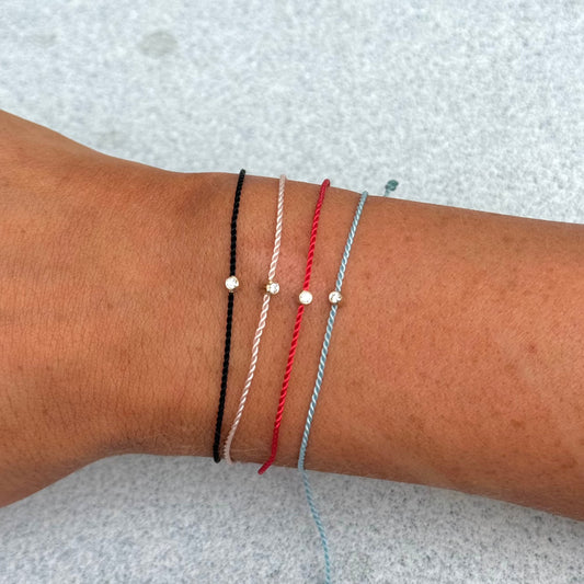 A wrist adorned with four thin braided bracelets in black, white, red, and blue, each featuring a small bead at the center. Among them, the SOL Gold Cord Bracelet by hortense stands out with its adjustable design and gleaming 14kt yellow gold charm.