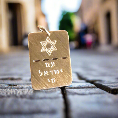 On a blurred cobblestone street, the BTH BRING THEM HOME Gold Dog Tag by Dylan James Jewelry/flower, intricately engraved with the Star of David and Hebrew letters, rests elegantly.