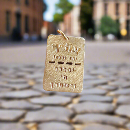 The BTH BRING THEM HOME Gold Dog Tag by Dylan James Jewelry/flower, featuring intricate Hebrew letters and delicate engravings, elegantly hangs against a blurred cobblestone street backdrop.